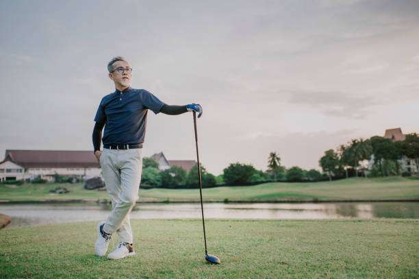 Senior Asian man smiling, leaning on his golf club on a beautiful course.