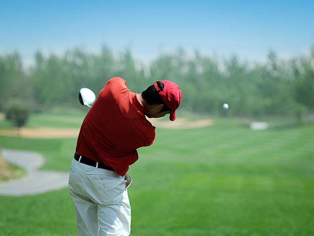 Golfer in red shirt swinging a golf club, hitting the ball on a green course.