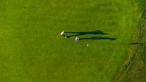 Aerial view: two golfers and their shadows on a green, near a golf club bag.