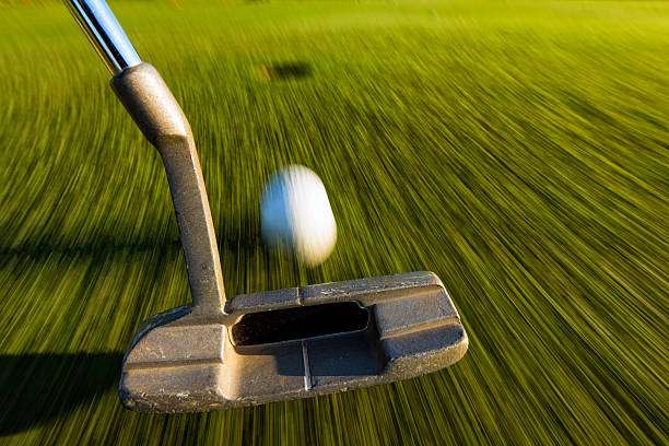 Close-up of a putter golf club striking a golf ball on a blurred green.