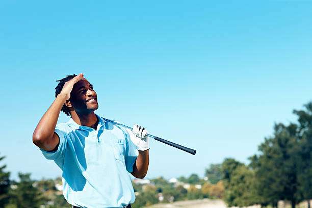 Smiling male golfer holding a golf club, shielding his eyes from the bright sun.